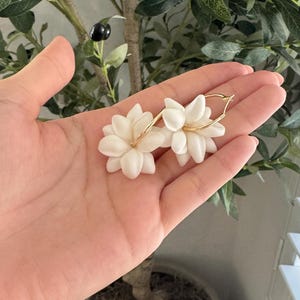 May include: White flower earrings with gold hoops. The earrings are in the shape of a flower with multiple petals. The earrings are held in a hand, with a plant in the background.