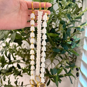 Pu&ograve; includere: Tre cordini bianchi con perline e accessori color oro. Ogni cordino presenta un motivo ripetuto di perline bianche a forma di fiore e piccole perline dorate. I cordini hanno chiusure e anelli color oro.
