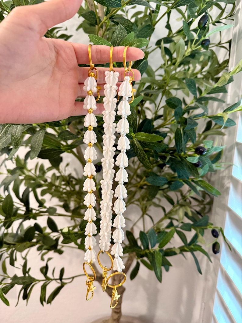 Pu&ograve; includere: Tre cordini bianchi con perline e accessori color oro. Ogni cordino presenta un motivo ripetuto di perline bianche a forma di fiore e piccole perline dorate. I cordini hanno chiusure e anelli color oro.
