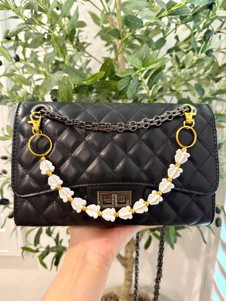 Pu&ograve; includere: Borsa nera trapuntata con tracolla a catena e decorativi accenti floreali bianchi. La borsa presenta finiture dorate e una chiusura rettangolare. La borsa &egrave; tenuta da una mano.