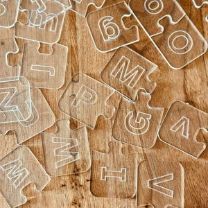 May include: Clear acrylic puzzle pieces with etched letters and numbers scattered on a wooden surface. The pieces are transparent with white outlines, featuring the letters M, P, V, and numbers like 6. The wooden surface has a warm, brown tone.