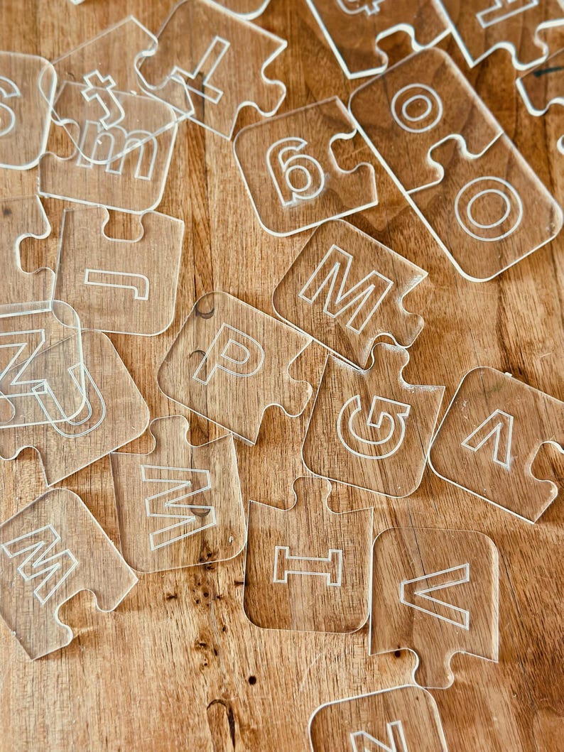 May include: Clear acrylic puzzle pieces with etched letters and numbers scattered on a wooden surface. The pieces are transparent with white outlines, featuring the letters M, P, V, and numbers like 6. The wooden surface has a warm, brown tone.