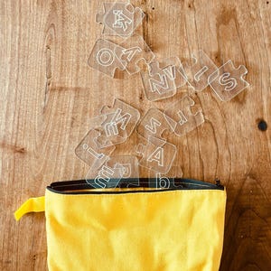 May include: A yellow zippered pouch with a black zipper and a yellow pull tab. Clear puzzle pieces with white letters are scattered above the pouch, resting on a wooden surface.