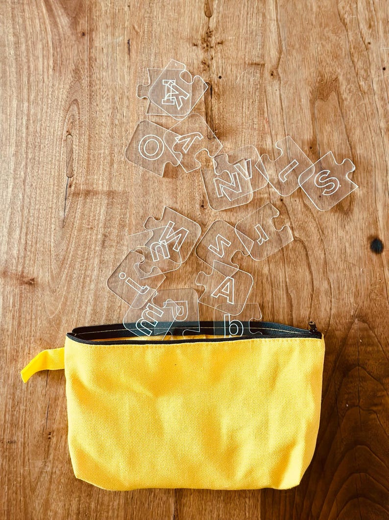 May include: A yellow zippered pouch with a black zipper and a yellow pull tab. Clear puzzle pieces with white letters are scattered above the pouch, resting on a wooden surface.