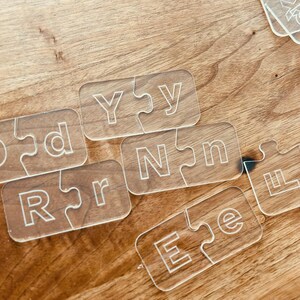 May include: Clear acrylic puzzle pieces with white engraved letters spelling out "DADDY" and "LOVE" on a wooden surface. The pieces are interlocking, creating a fun and educational toy.