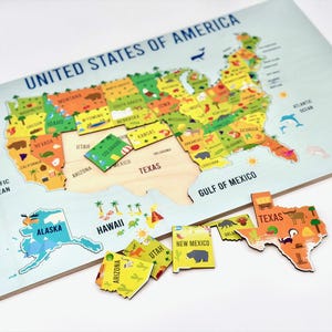 May include: Wooden United States map puzzle with colorful state pieces. The puzzle features the text "UNITED STATES OF AMERICA" and includes state names and illustrations. Some state pieces are removed, showing the wooden base.