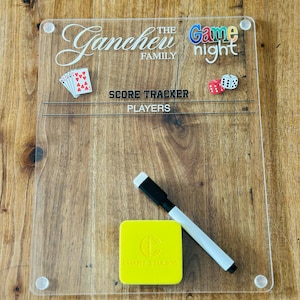 Könnte beinhalten: Transparente Acryl-Spieltafel mit dem Text "The Ganchev Family Game Night". Enthält ein Kartenspiel, Würfel, einen gelben Radierer und einen Whiteboard-Marker. Die Tafel liegt auf einer Holzoberfläche.