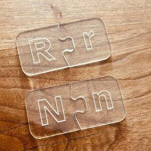 May include: Two clear acrylic puzzle pieces with the letters "R" and "r" etched into them, and two more pieces with "N" and "n". The pieces are on a wooden surface.