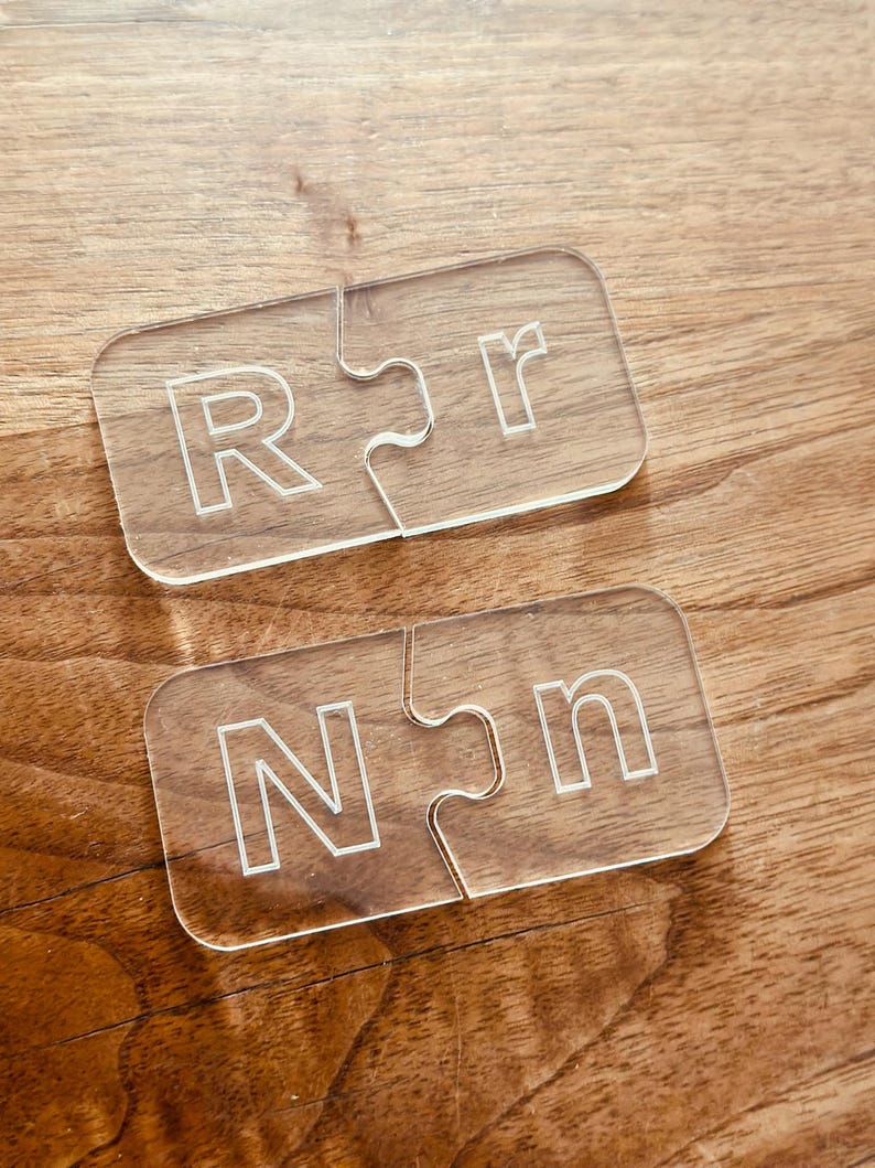 May include: Two clear acrylic puzzle pieces with the letters "R" and "r" etched into them, and two more pieces with "N" and "n". The pieces are on a wooden surface.