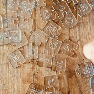 May include: Clear acrylic puzzle pieces scattered on a wooden surface. Each piece features a white letter or number. The pieces are interlocking, suggesting a learning or educational toy.