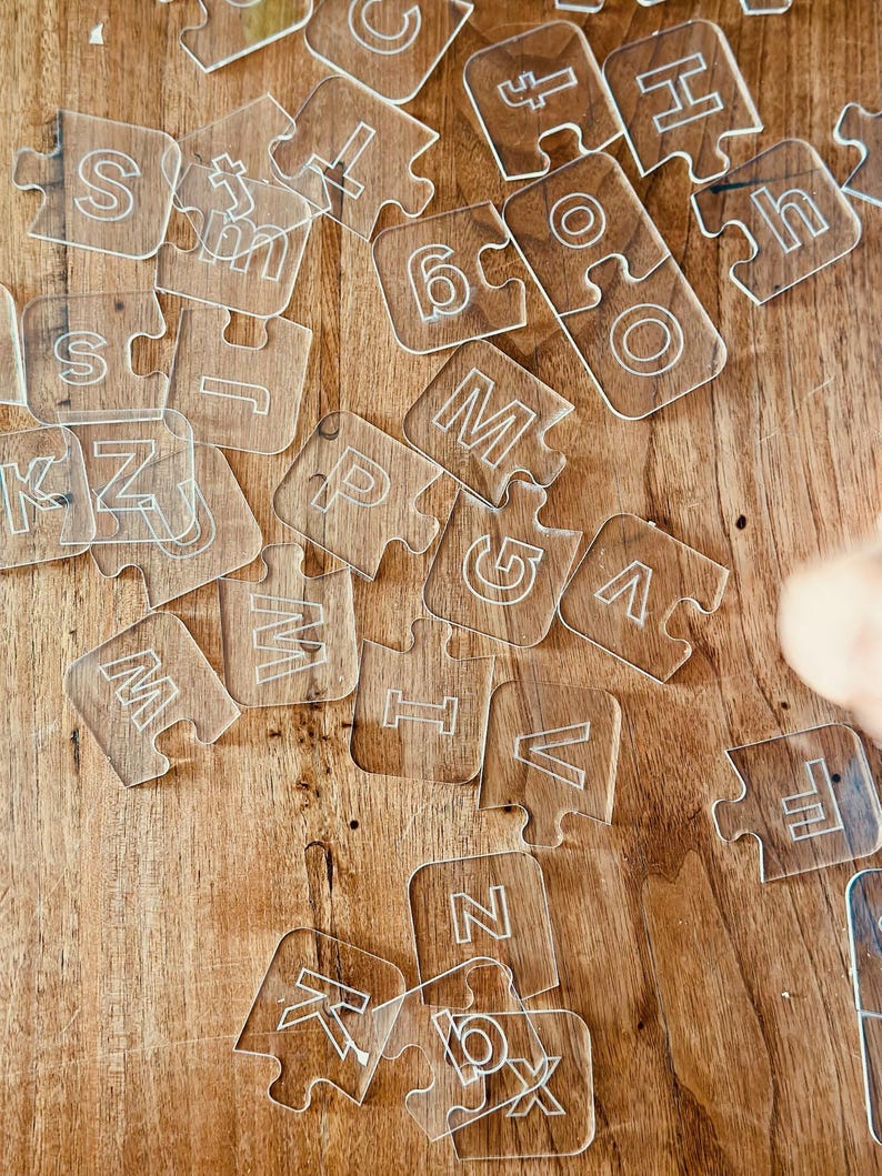 May include: Clear acrylic puzzle pieces scattered on a wooden surface. Each piece features a white letter or number. The pieces are interlocking, suggesting a learning or educational toy.