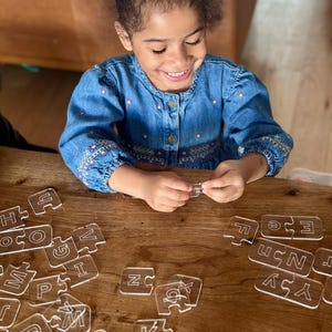May include: A young person smiles while assembling a clear acrylic alphabet puzzle on a wooden table. The puzzle pieces, each shaped like a letter, are scattered around. The person is wearing a blue denim dress.