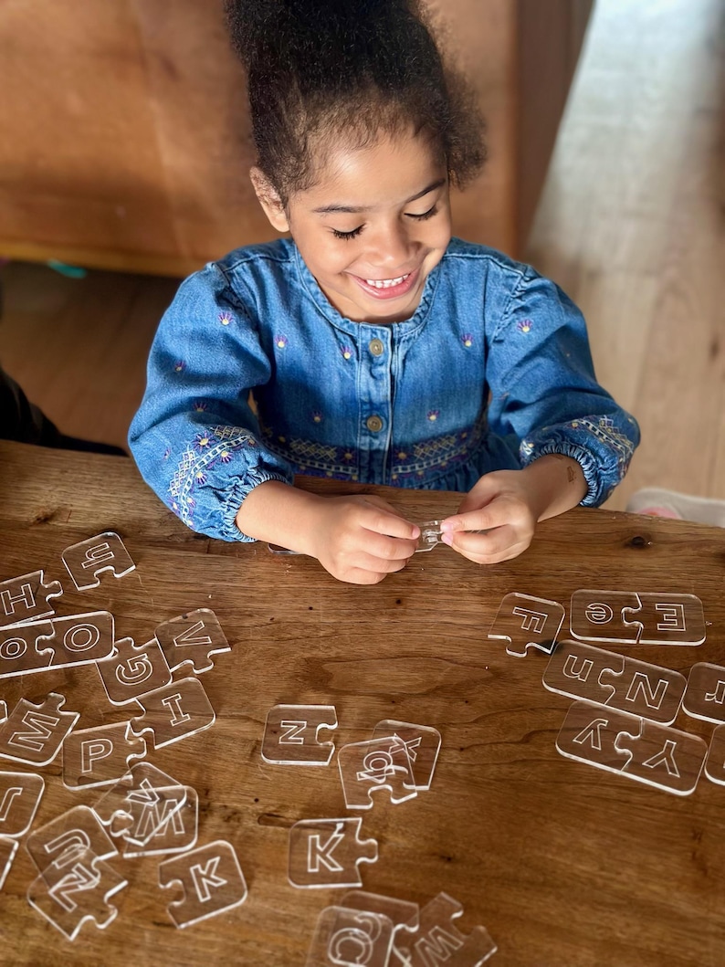 May include: A young person smiles while assembling a clear acrylic alphabet puzzle on a wooden table. The puzzle pieces, each shaped like a letter, are scattered around. The person is wearing a blue denim dress.