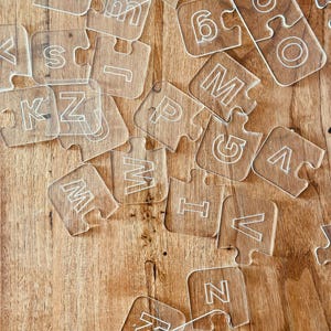 May include: Clear acrylic puzzle pieces scattered on a wooden surface. Each piece features a white letter or number, including the letters S, K, M, and Z, and the number 6. The pieces are interlocking, suggesting a learning or educational theme.