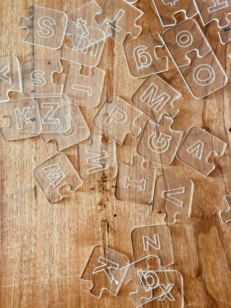 May include: Clear acrylic puzzle pieces scattered on a wooden surface. Each piece features a white letter or number, including the letters S, K, M, and Z, and the number 6. The pieces are interlocking, suggesting a learning or educational theme.
