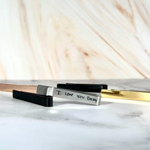 May include: A collection of metal tie clips in various colors, including gold, silver, black, and rose gold. One clip is inscribed with "I love you Baby". The clips are rectangular and lie on a marble surface.