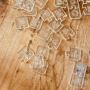 May include: Clear acrylic puzzle pieces scattered on a wooden surface. Each piece is shaped like a jigsaw puzzle and features a letter or number. The letters and numbers are etched into the clear acrylic.