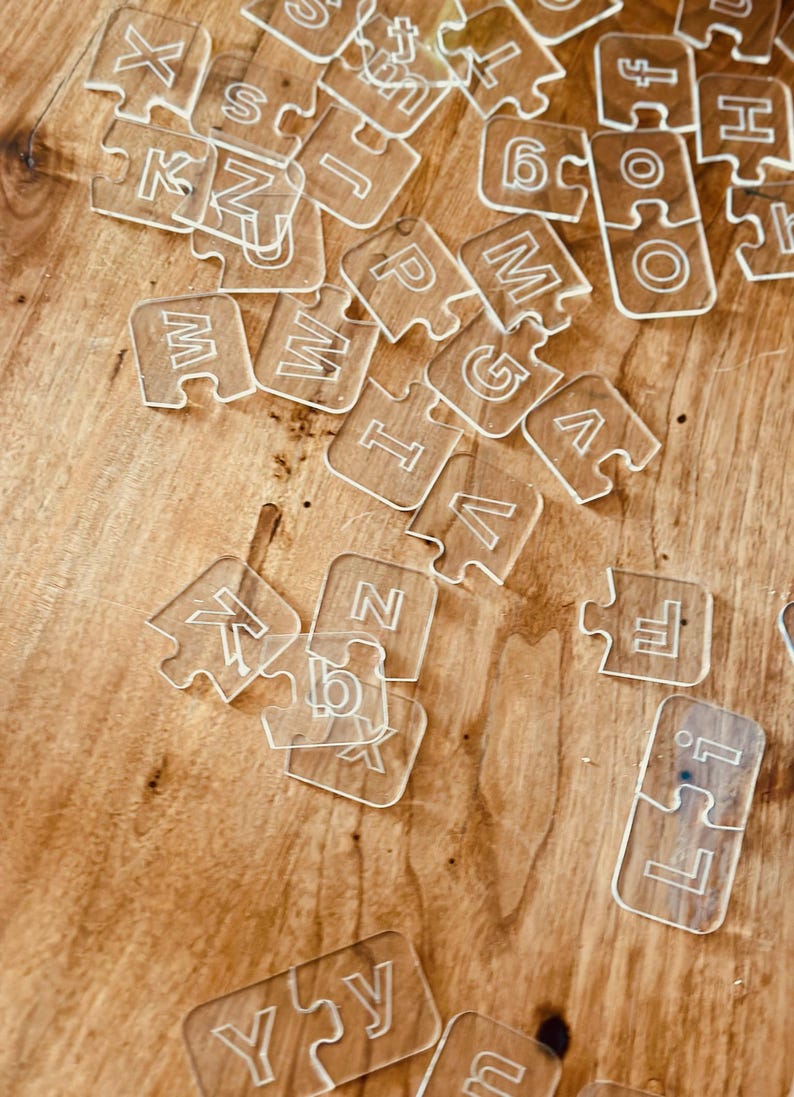 May include: Clear acrylic puzzle pieces scattered on a wooden surface. Each piece is shaped like a jigsaw puzzle and features a letter or number. The letters and numbers are etched into the clear acrylic.