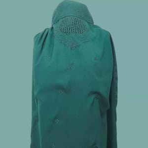 May include: A teal green long-sleeved shirt with a decorative embroidered design on the back. The design features a circle with a grid pattern and a floral motif.