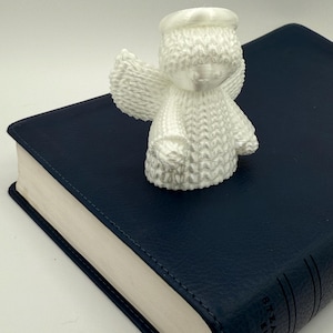 Puede incluir: Figurita de &aacute;ngel de cer&aacute;mica blanca con halo, alas y superficie texturizada. El &aacute;ngel est&aacute; sentado sobre un libro encuadernado en cuero azul.