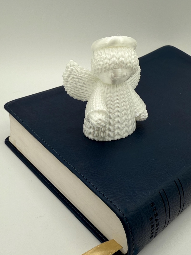 Puede incluir: Figurita de &aacute;ngel de cer&aacute;mica blanca con halo, alas y superficie texturizada. El &aacute;ngel est&aacute; sentado sobre un libro encuadernado en cuero azul.