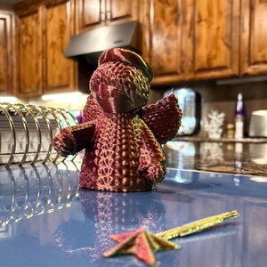 Puede incluir: Una peque&ntilde;a figurita de &aacute;ngel impresa en 3D con una superficie texturizada. El &aacute;ngel es de color burdeos oscuro y tiene un halo. El &aacute;ngel est&aacute; sentado sobre una superficie azul.