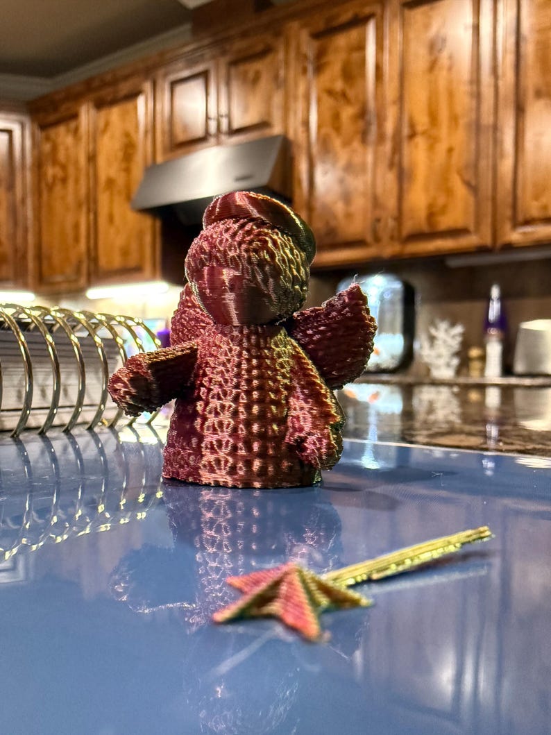 Puede incluir: Una peque&ntilde;a figurita de &aacute;ngel impresa en 3D con una superficie texturizada. El &aacute;ngel es de color burdeos oscuro y tiene un halo. El &aacute;ngel est&aacute; sentado sobre una superficie azul.