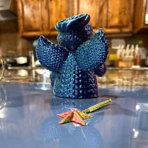 Puede incluir: Una figurita de &aacute;ngel azul impresa en 3D con un halo y alas. El &aacute;ngel est&aacute; de pie sobre una superficie azul con un objeto en forma de estrella dorada y rosa delante de &eacute;l.