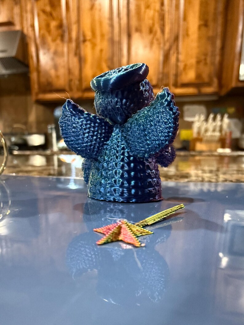 Puede incluir: Una figurita de &aacute;ngel azul impresa en 3D con un halo y alas. El &aacute;ngel est&aacute; de pie sobre una superficie azul con un objeto en forma de estrella dorada y rosa delante de &eacute;l.