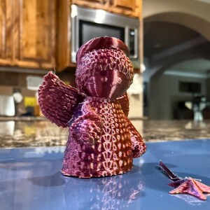 Puede incluir: Una figurita de &aacute;ngel impresa en 3D con un degradado de color rosa y morado. El &aacute;ngel tiene un dise&ntilde;o detallado con una superficie texturizada. A la figurita le falta un ala.