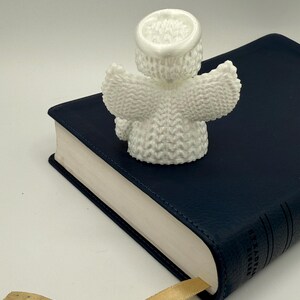 Puede incluir: Figurita de &aacute;ngel de punto blanco con alas y halo, sentada sobre un libro encuadernado en cuero azul con un marcap&aacute;ginas de cinta dorada.