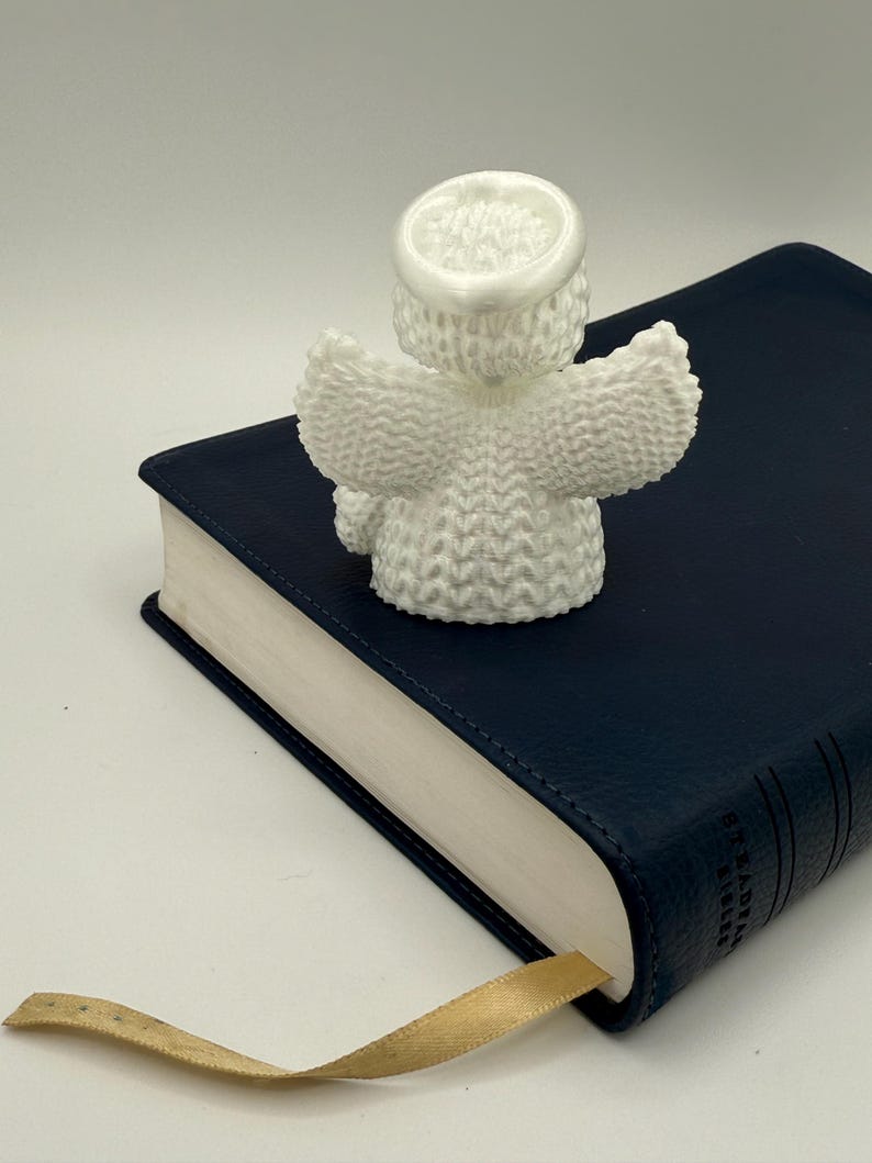 Puede incluir: Figurita de &aacute;ngel de punto blanco con alas y halo, sentada sobre un libro encuadernado en cuero azul con un marcap&aacute;ginas de cinta dorada.