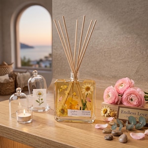 May include: A square glass reed diffuser filled with yellow liquid and pressed flowers, featuring a white label with text. It includes wooden reeds, a small candle in a glass cloche, and pink ranunculus flowers on a wooden surface.