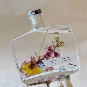 Könnte beinhalten: Eine klare, quadratische Reed-Diffusor-Flasche mit silbernem Verschluss, gefüllt mit Flüssigkeit und bunten Trockenblumen. Das Etikett lautet "Cashmere Plum" und "Reed Diffuser 350ml". Die Flasche steht auf einer hellen, strukturierten Oberfläche.