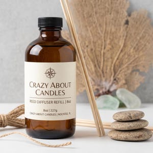 Könnte beinhalten: Eine bernsteinfarbene Glasflasche mit schwarzem Verschluss, die Nachfüllflüssigkeit für einen Raumduft enthält. Das weiße Etikett trägt die Aufschrift Crazy About Candles, Reed Diffuser Refill, 8oz, 227g und Crazy About Candles, Nocatee, FL.