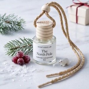 May include: A clear glass 8ml car diffuser bottle with a light wood cap featuring the text SCENT LOGIC LAB. The bottle label reads SCENT LOGIC LAB The North Pole SCENTED CAR DIFFUSER 8ml. It hangs from a tan cord with gold metallic threading.