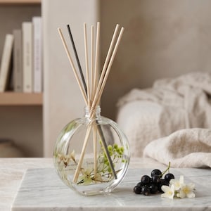 May include: A clear, round glass reed diffuser bottle containing liquid and small dried flowers. Several light-coloured wooden reeds and one dark grey reed are inserted into the neck. A small cluster of fresh blackcurrants and white flowers sits nearby.