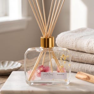May include: A clear, square glass reed diffuser with a gold-toned collar, containing pink dried flowers and light-colored reeds. A white rectangular label on the front features the printed text White Orris and Reed Diffuser Oil.