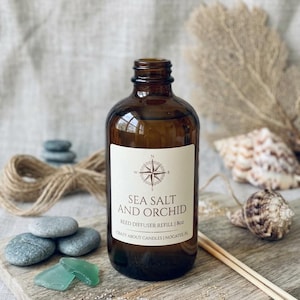 Puede incluir: Una botella de vidrio ámbar que contiene aceite de recarga para difusor de varillas. La etiqueta blanca presenta un gráfico de rosa de los vientos y el texto: SEA SALT AND ORCHID, REED DIFFUSER REFILL | 8oz, CRAZY ABOUT CANDLES | NOCATEE, FL.