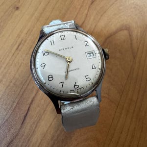 Puede incluir: Un reloj de pulsera Kienzle vintage con caja plateada y esfera blanca. El reloj presenta números negros, una ventana de fecha y una correa de cuero gris claro. La esfera del reloj dice "Kienzle" y "Magnetic".