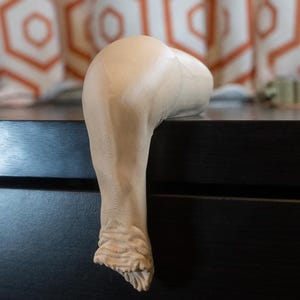 May include: A close-up of a beige, textured, fabric leg form. The leg form is bent at the knee and the foot is curled under. The leg form is resting on a dark brown surface.