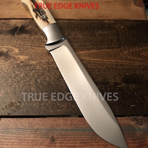 Puede incluir: Un cuchillo grande de acero inoxidable con una hoja pulida y un mango de hueso y metal. El texto "TRUE EDGE KNIVES" está impreso en rojo. El cuchillo se muestra sobre una superficie de madera.
