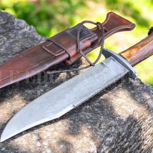 Puede incluir: Un cuchillo de caza grande con una hoja de acero pulido y un mango de madera descansa sobre una superficie de piedra desgastada. El cuchillo va acompañado de una funda de cuero marrón. El mango tiene un aspecto natural y texturizado.