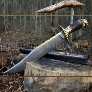 May include: A large hunting knife with a curved blade and a handle made of antler and brass. The blade is silver with a gold accent along the spine. The knife rests on a wooden block, with a small folding knife nearby.