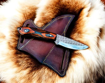 May include: A brown leather sheath with a stitched edge holds a small knife with a wood handle. The handle has a dark blue and red wood grain pattern. The blade is made of steel and has a serrated edge.