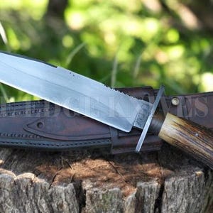 Peut inclure: Un grand couteau de chasse avec une lame argentée et un manche en corne marron repose sur un étui en cuir. Le couteau a un bord dentelé près du manche. L'étui est marron et présente des coutures décoratives.