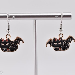 Puede incluir: Un par de pendientes con forma de murciélago. Cada pendiente presenta un murciélago negro con cara blanca, cuernos rojos y alas doradas. Los pendientes están suspendidos de ganchos plateados. Se exhiben en un soporte plateado.