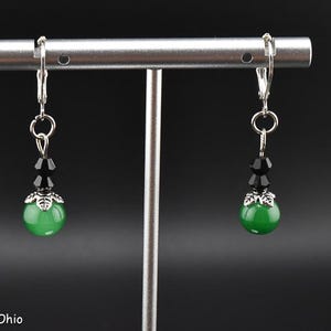 May include: A pair of dangle earrings with lever-back closures. Each earring features a green, round bead, accented with black beads and a silver-colored decorative cap. The earrings are displayed on a silver-colored stand against a dark background. The text "Abbys Accessories Ohio" is visible.