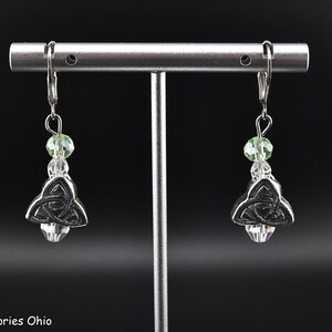 May include: A pair of silver-toned Celtic knot earrings with green and clear crystal accents. The earrings are displayed on a silver stand against a black background. The text "Abbys Accessories Ohio" is visible at the bottom.