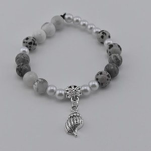 May include: A gray and white beaded bracelet with a silver seashell charm. The bracelet is made with white and gray speckled beads, white pearls, and silver beads.